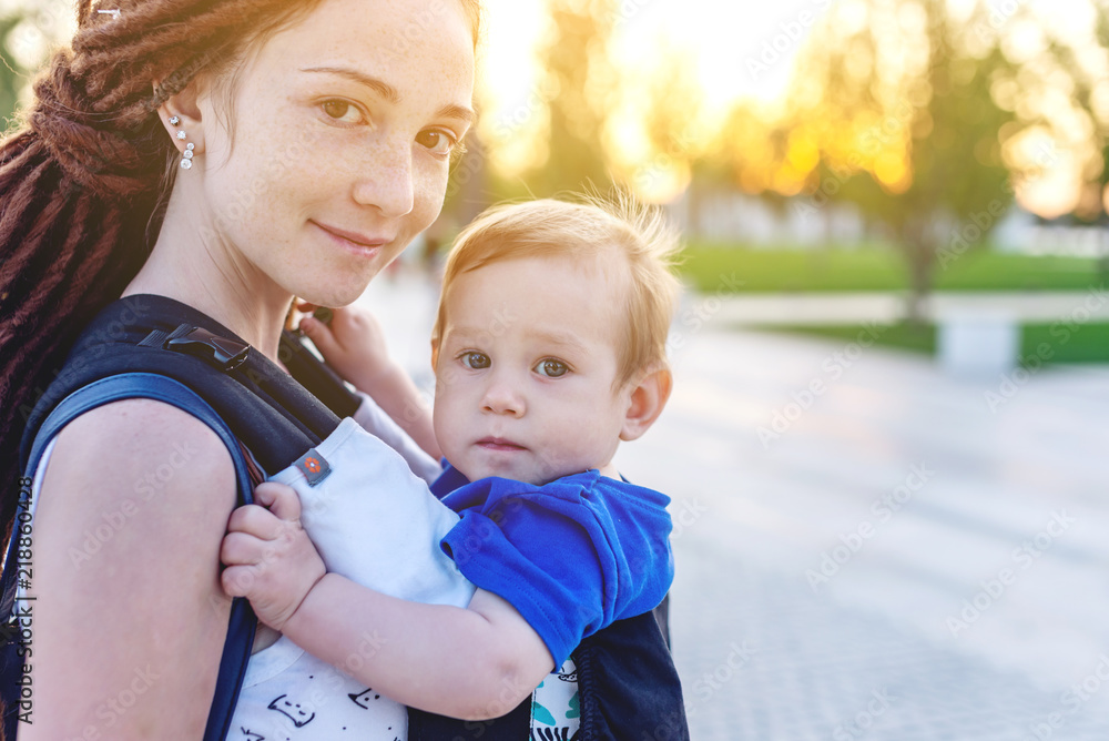 Young happy mom with baby son in ergo backpack travel together. Sunny summer day in the Park. Concept of modern parents Young happy mom with baby son in ergo backpack travel together. Sunny summer day in the Park. Concept of modern parents