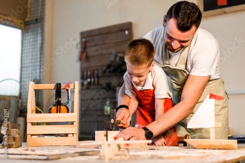 Dad teaches his son carpentry in the workshop playing hammering nails into a wooden bar close-up