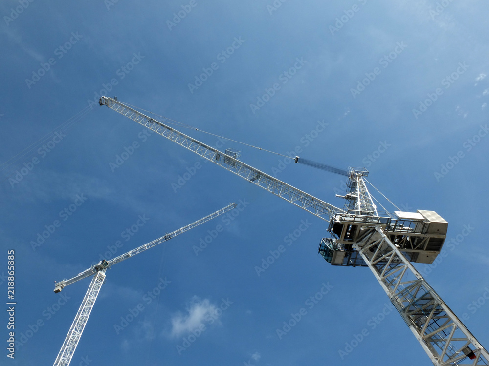 looking upward view of two tall white construction cranes on a building site against a blue sky with clouds looking upward view of two tall white construction cranes on a building site against a blue sky with clouds