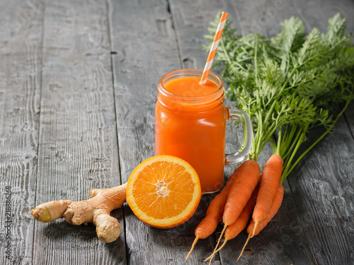 A mug of fresh carrot smoothie with cocktail straw, parsley, carrots, ginger root and oranges on a table.