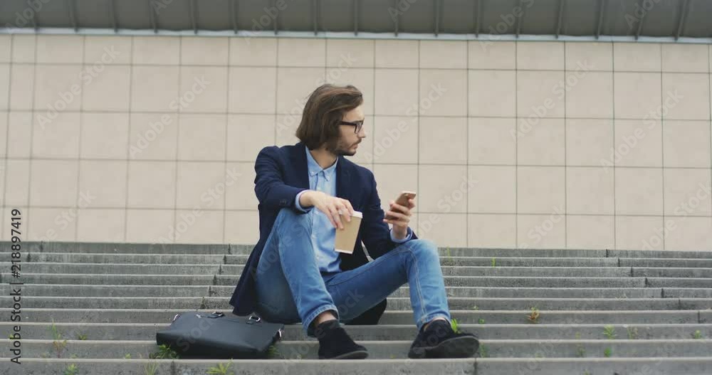 Caucasian stylish young man looking at the sides while sipping coffee, sitting on steps in the city center and holding smartphone. Caucasian stylish young man looking at the sides while sipping coffee, sitting on steps in the city center and holding smartphone.