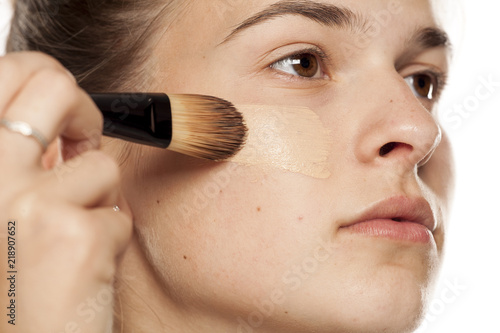 Young woman applying liquid foundation with brush on white background