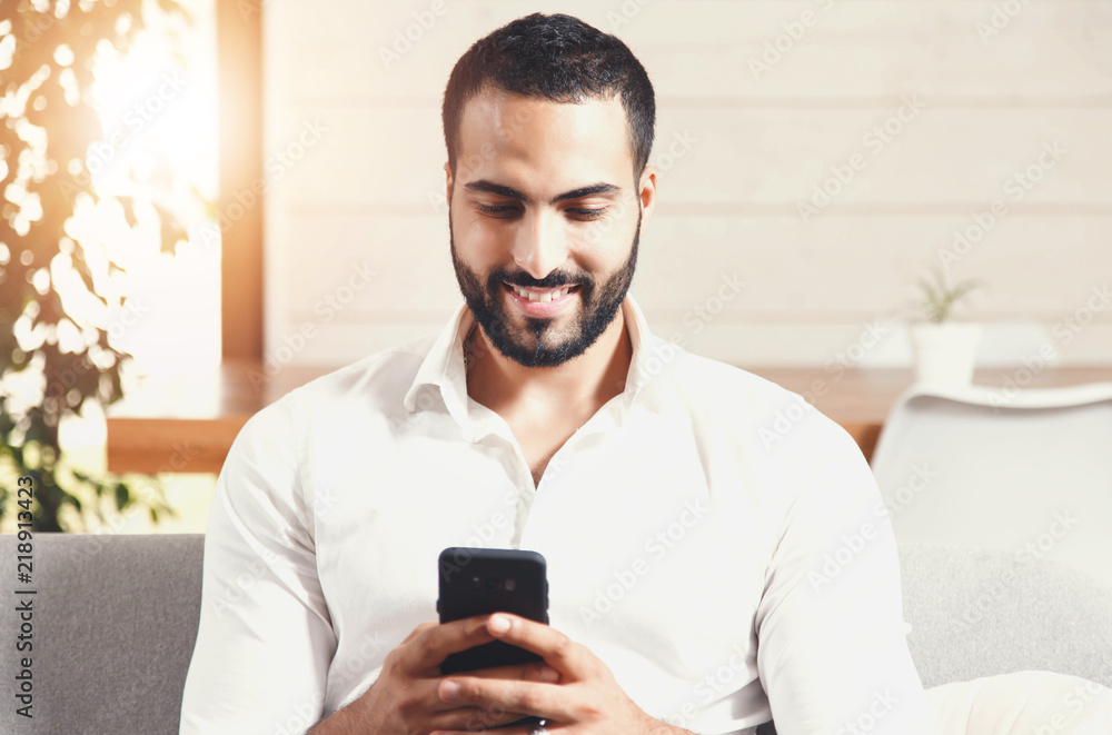 Sexy bearded man in white shirt sitting on the sofa and writting message on the smartphone, using app concept Sexy bearded man in white shirt sitting on the sofa and writting message on the smartphone, using app concept