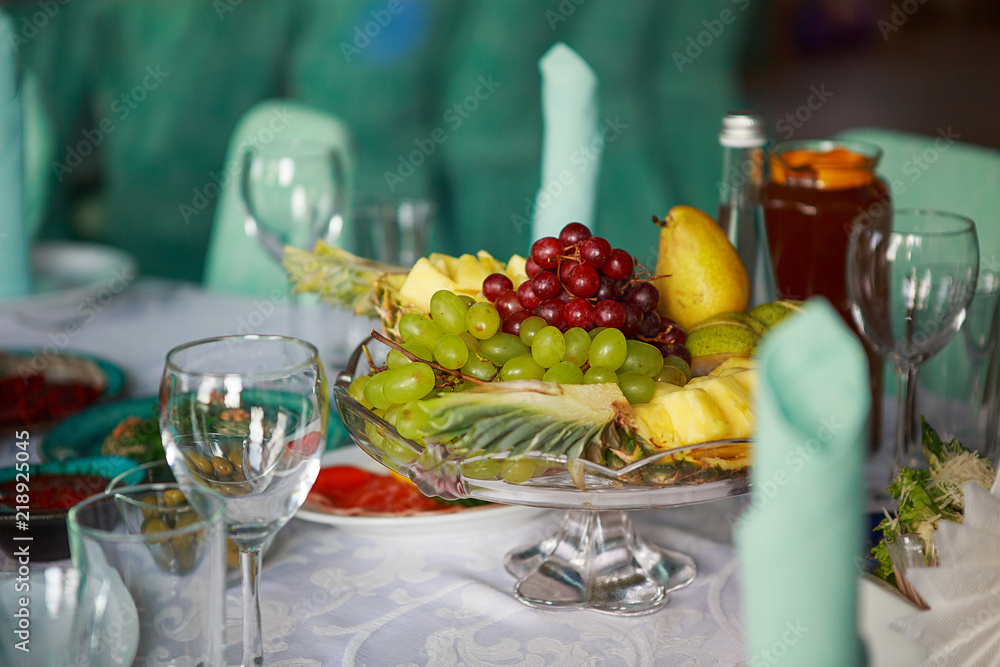 a still-life of exotic fruit. On a glass dish, grapes, pineapple, pear. The table is set for a party