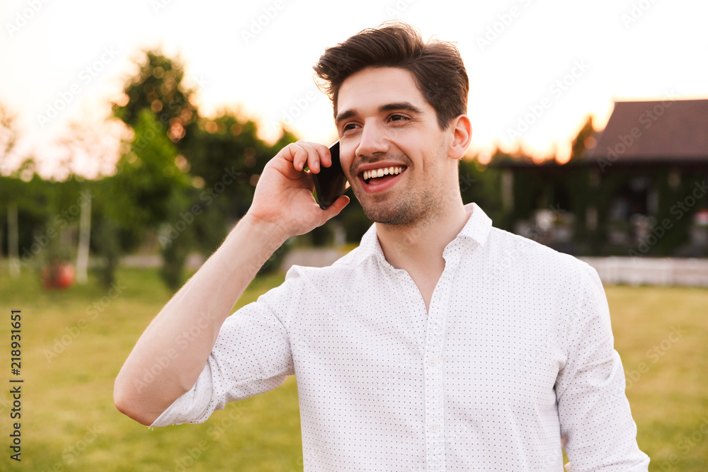 Image of caucasian happy man 25-30 wearing white shirt smiling, and talking on black smartphone while walking outdoor on nature Image of caucasian happy man 25-30 wearing white shirt smiling, and talking on black smartphone while walking outdoor on nature