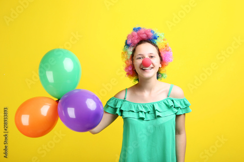 Young girl in clown wig and balloons on yellow background