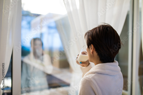 Rear view of Asian woman drinking a glass of milk in living room and looking outside window in the morning