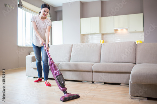 A picture of woman stands in studio apartment and cleaning the floor. She uses vacuum cleaner for that. Girl is looking down.