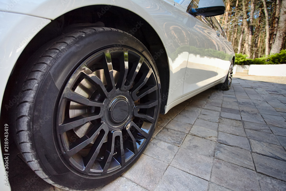 Modern auto wheel of car with beautiful black alloy disc and fresh rubber tread on paving slab with green pines on sunset background