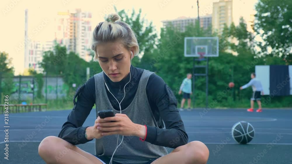 Beautiful young woman sitting on basketball court and typing on phone Beautiful young woman sitting on basketball court and typing on phone