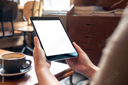 Mockup image of woman's hands holding black tablet pc with blank white screen with coffee cup on wooden table in cafe
