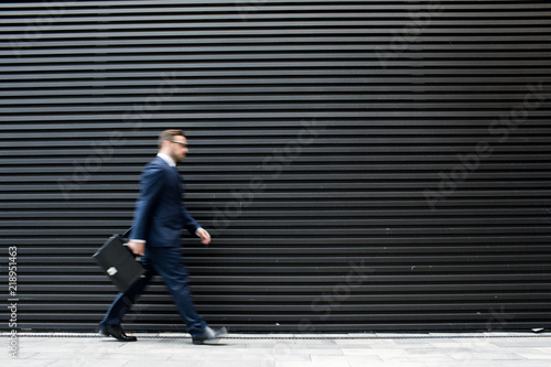 Side view of young successful blurred male in business dress, eyeglasses and briefcase going near black profiled sheeting 