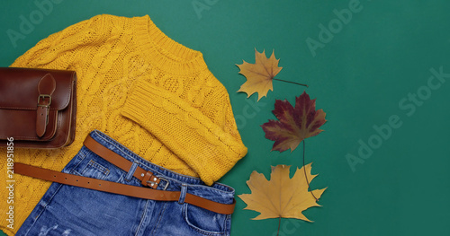 Female orange knitted sweater, blue jeans, leather bag and autumn leaves on green background top view flat lay. Fashion Lady Clothes Set Trendy Cozy Knit Jumper Autumn accessories Female fashion look
