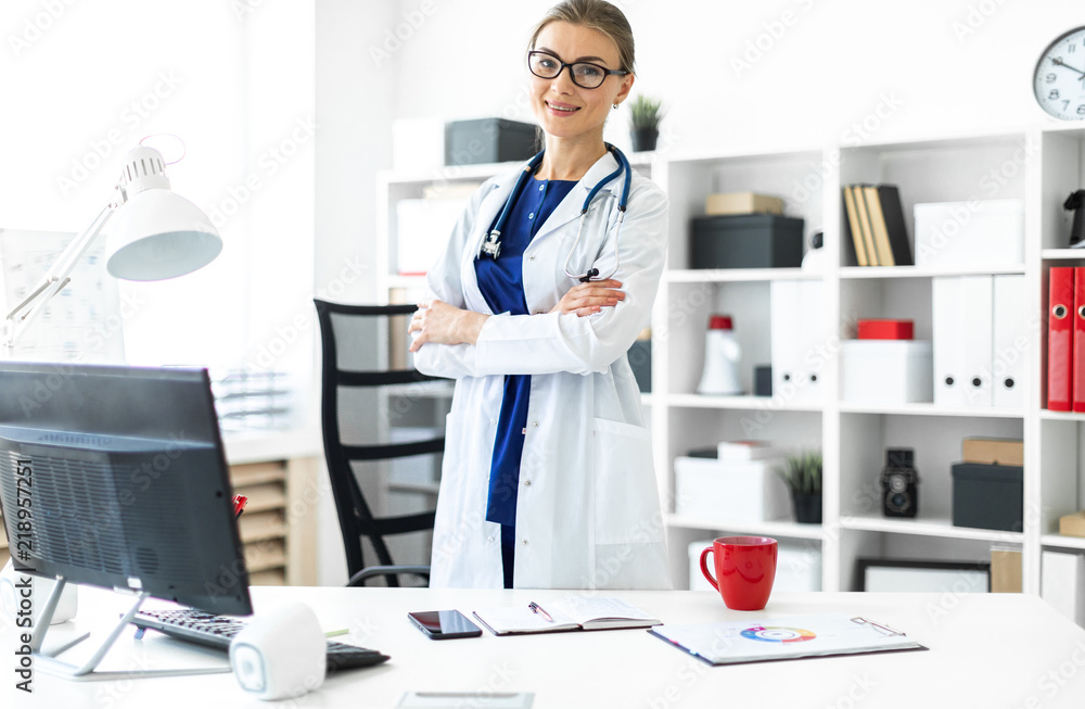 A young girl in a white coat is standing near a table in her office. A stethoscope hangs around her neck.