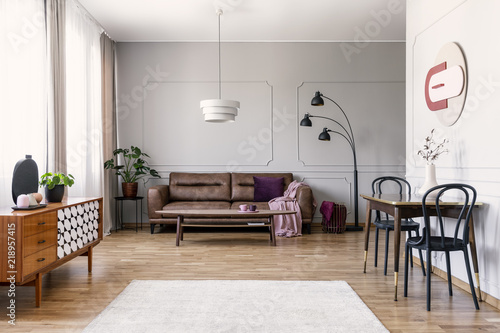 Real photo of light grey living room interior with window with curtains, leather couch, table with two chairs carpet on wooden floor and two lamps photo