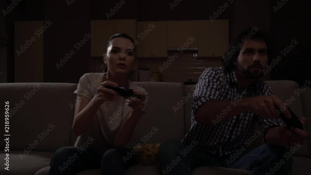 Man and woman sit on sofa and play game. They lean to the right together. People use joysticks. Then guy leans to the left. Girl repeats move in couple seconds. The are concentrated on game. Man and woman sit on sofa and play game. They lean to the right together. People use joysticks. Then guy leans to the left. Girl repeats move in couple seconds. The are concentrated on game.