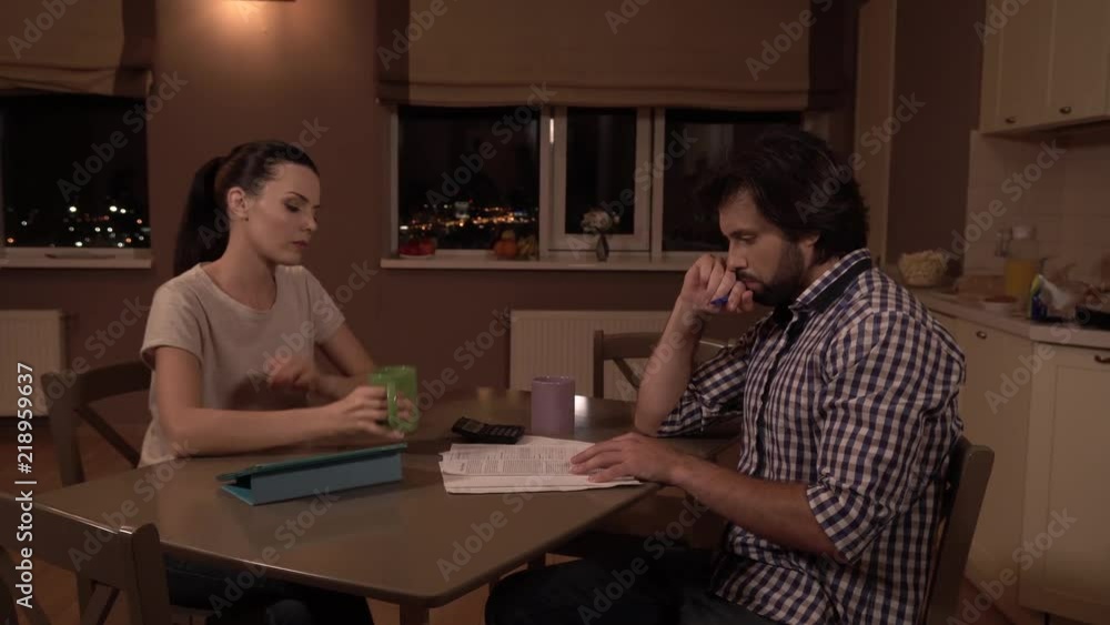 Upset man and woman sit on the table. They are thinking. Girl drinks from cup and looks at husband. He looks at papers and hold fist close to mouth. Man is thinking. They are worry. Upset man and woman sit on the table. They are thinking. Girl drinks from cup and looks at husband. He looks at papers and hold fist close to mouth. Man is thinking. They are worry.