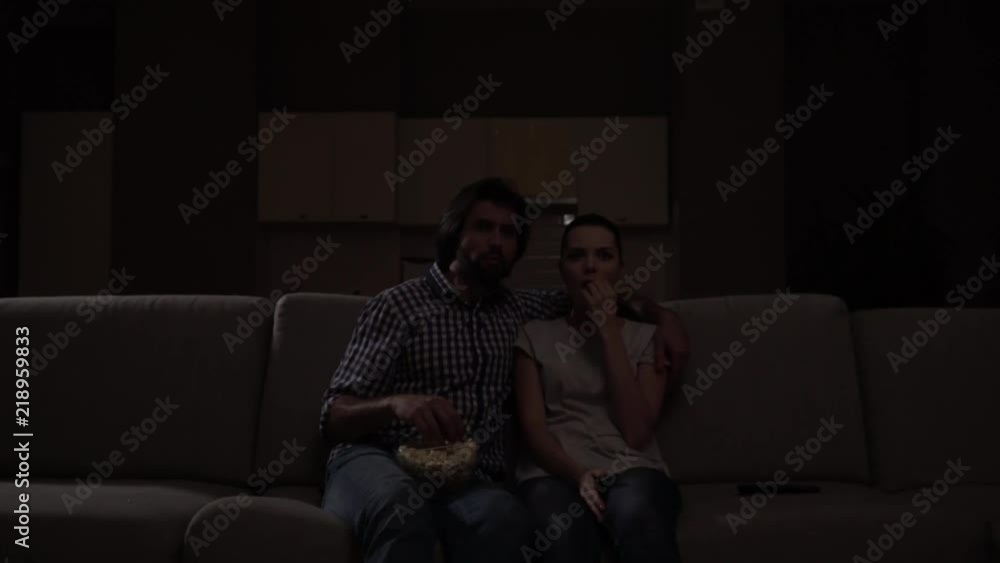 Man and woman sit on sofa and watching movie. They eat popcorn. Guy holds bowl with junk food on knees. Girl got scared. She coveres mouth with hand. Guy protects her. Man and woman sit on sofa and watching movie. They eat popcorn. Guy holds bowl with junk food on knees. Girl got scared. She coveres mouth with hand. Guy protects her.