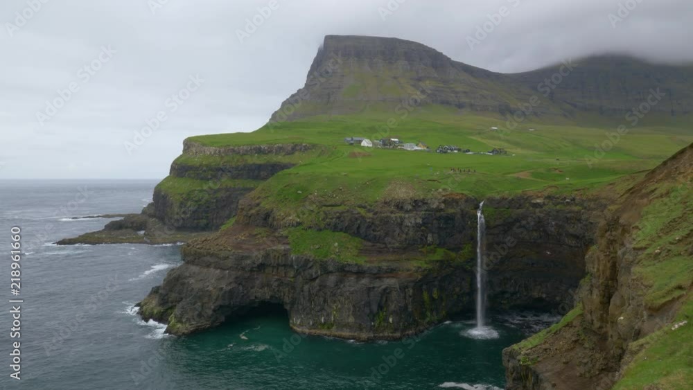 AERIAL: Beautiful waterfall near a quiet village in the green Scandinavian countryside flows off a rocky edge into the cold sea on cloudy morning. Breathtaking view of waterfall in rural Faroe Islands AERIAL: Beautiful waterfall near a quiet village in the green Scandinavian countryside flows off a rocky edge into the cold sea on cloudy morning. Breathtaking view of waterfall in rural Faroe Islands