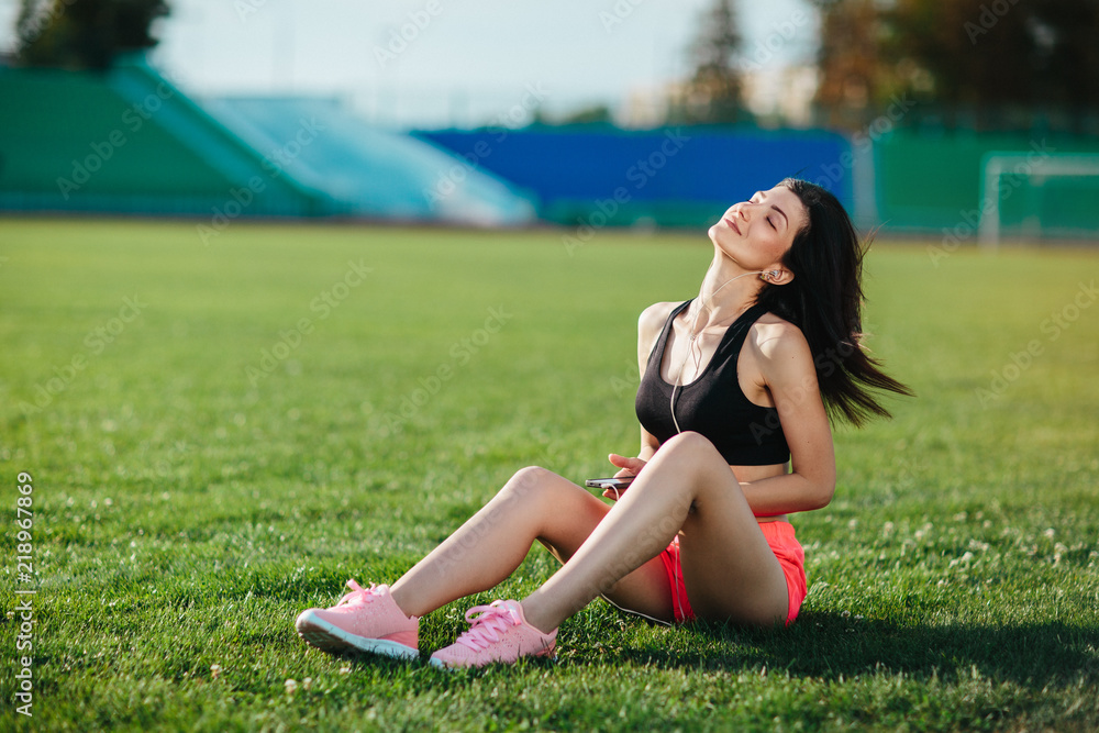 Young sporty joyful woman brunette in sport dress sitting on the grass football field stadium and listens to music in earphones, her head thrown back, she gets pleasure, relaxes