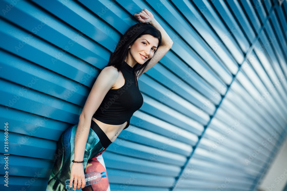Beautiful young fitness brunette girl in the top and the leggings with water bottle standing near blue wall looks at the camera and smiles