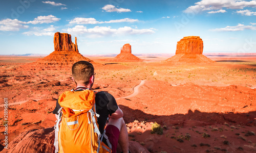 Monument Valley background. Amazed young traveler with backpack sits and absorbs the Navajo country desert landscape of famous large red rock domes on Utah-Arizona border, United States of America