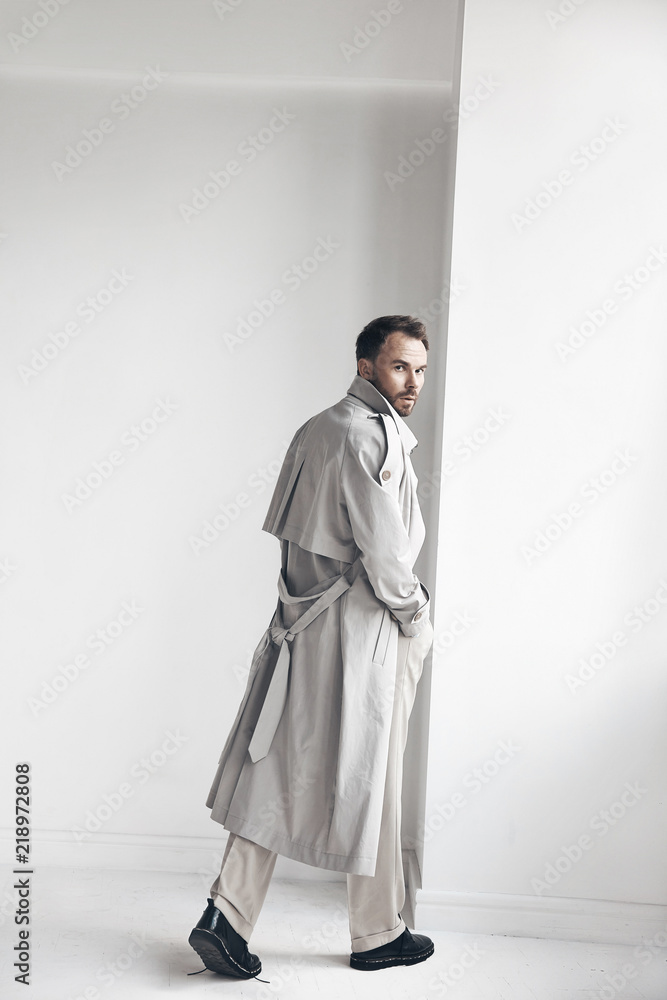 Fashion portrait of handsome man with dark beard and hair, weared in light trench coat, beige pants and black shoes. Man stands near white wall and looks back Fashion portrait of handsome man with dark beard and hair, weared in light trench coat, beige pants and black shoes. Man stands near white wall and looks back