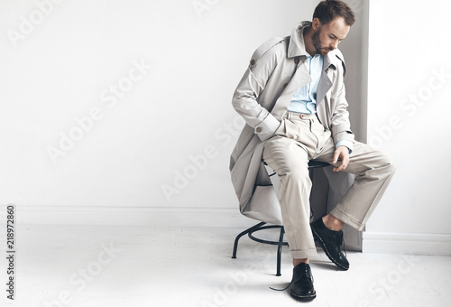 Fashion portrait of handsome  stylish man with dark beard and hair, weared in light trench coat, shirt, beige pants and black shoes. Man sits on the chair near white wall