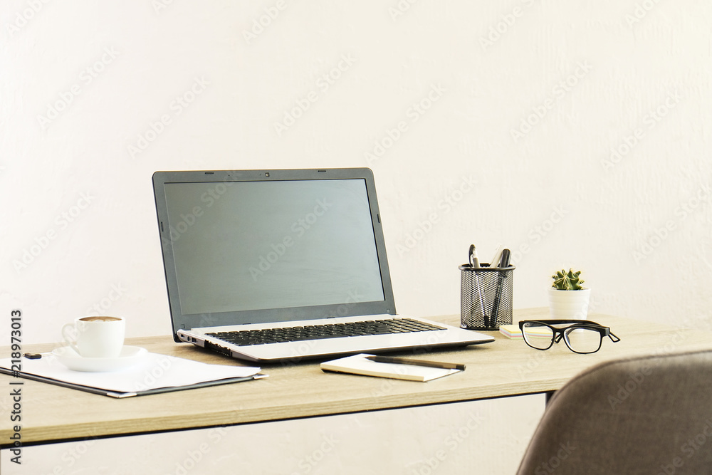 Blank screen laptop computer, cup of cappuccino coffee, cactus, supplies and folded eye glasses on wooden desk in spacious office full of sunlight. Creative workspace. Close up, copy space, background Blank screen laptop computer, cup of cappuccino coffee, cactus, supplies and folded eye glasses on wooden desk in spacious office full of sunlight. Creative workspace. Close up, copy space, background