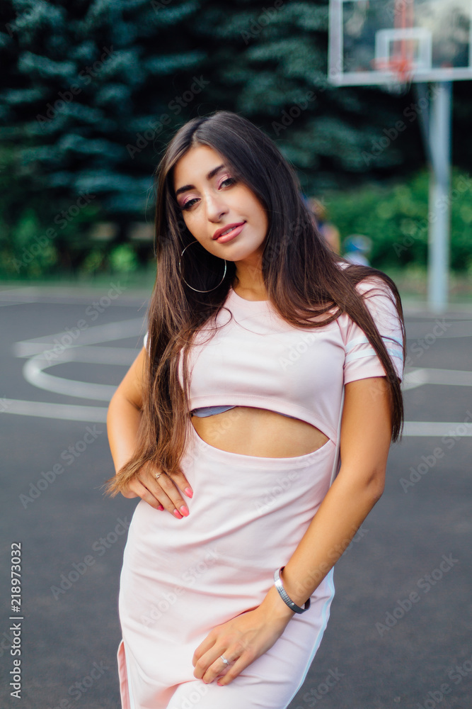 Charming brunette female dressed in a pink dress posing on the basketball court.