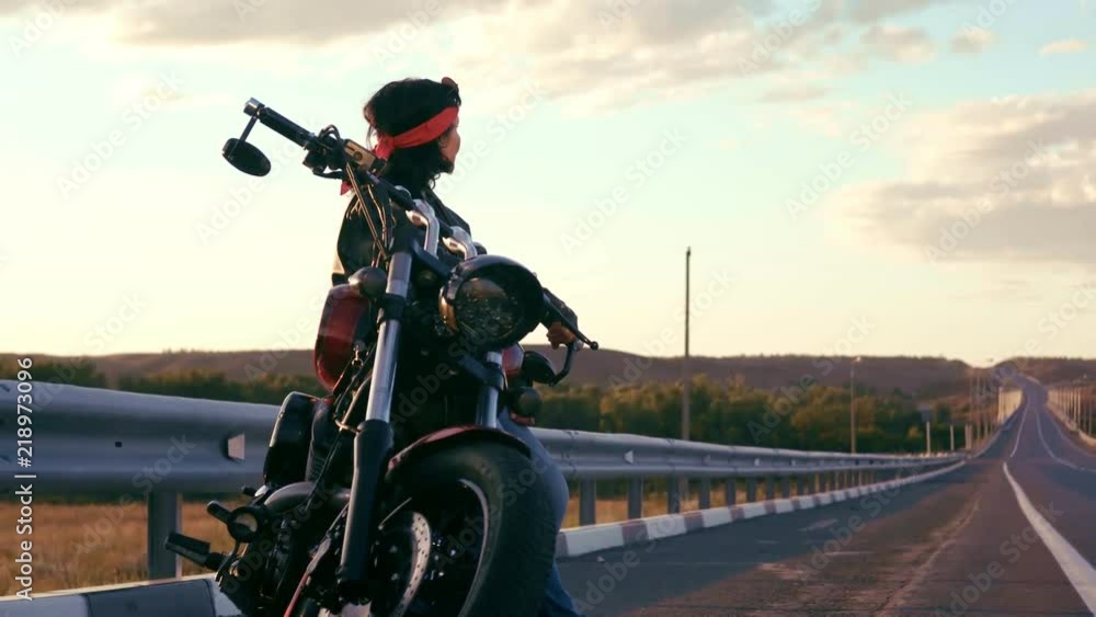 An elderly woman in a red bandana, leather jacket and gloves stands leaning on a steep motorcycle, against an empty highway. Hands folded on his chest, looks around. Happy life of an elderly rocker An elderly woman in a red bandana, leather jacket and gloves stands leaning on a steep motorcycle, against an empty highway. Hands folded on his chest, looks around. Happy life of an elderly rocker