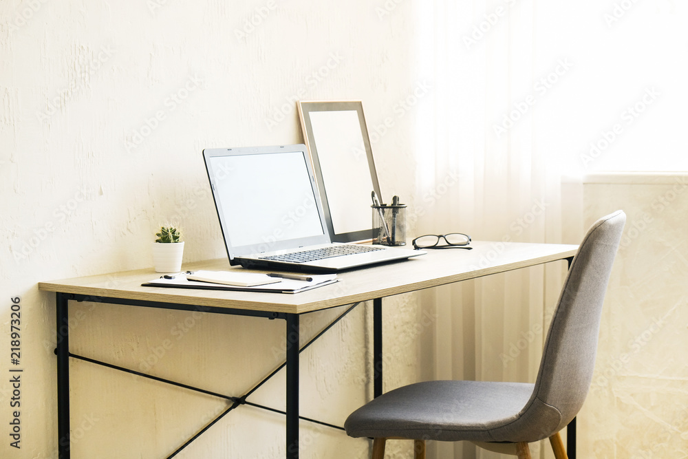 Laptop computer, clipboard, cactus home plant, supplies and folded glasses on wooden desk, gray chair in office full of sunlight. Designers creative workspace concept. Close up, copy space, background Laptop computer, clipboard, cactus home plant, supplies and folded glasses on wooden desk, gray chair in office full of sunlight. Designers creative workspace concept. Close up, copy space, background
