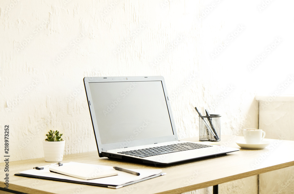 Blank screen laptop computer, cup of cappuccino coffee, cactus, supplies and folded eye glasses on wooden desk in spacious office full of sunlight. Creative workspace. Close up, copy space, background Blank screen laptop computer, cup of cappuccino coffee, cactus, supplies and folded eye glasses on wooden desk in spacious office full of sunlight. Creative workspace. Close up, copy space, background