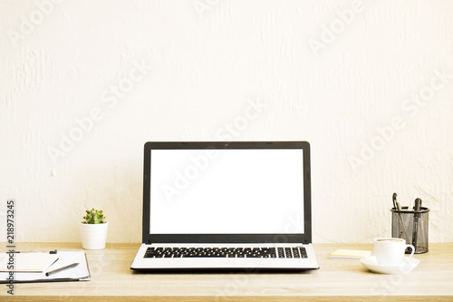Blank screen laptop computer, cup of cappuccino coffee, cactus, supplies and folded eye glasses on wooden desk in spacious office full of sunlight. Creative workspace. Close up, copy space, background
