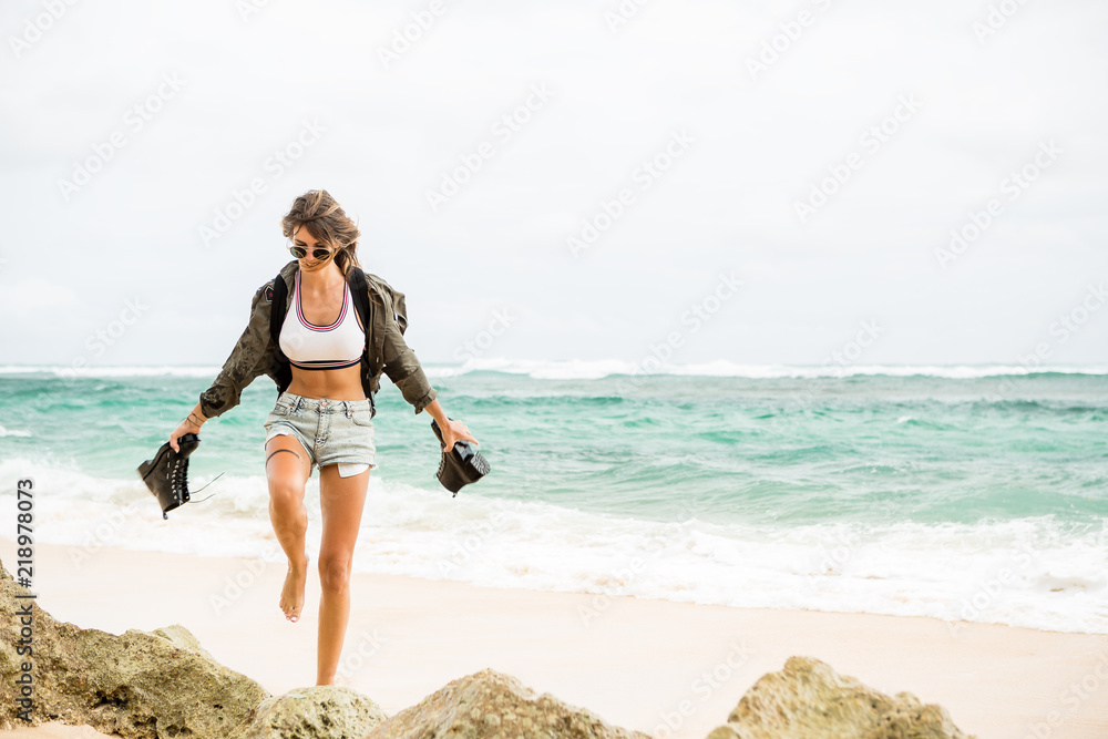 Hipster fit girl in sexy shorts, khaki shirt, sunglasses and travel backpack pour out water from army boots. Modern tourist traveler, active lifestyle, sport adventure camp and extreme on vacation. Hipster fit girl in sexy shorts, khaki shirt, sunglasses and travel backpack pour out water from army boots. Modern tourist traveler, active lifestyle, sport adventure camp and extreme on vacation.