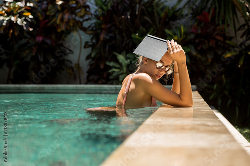 Beautiful fit woman in sexy bikini and sunglasses in a pool spa hotel make shadow with a note book. Relax vacation concept. Hot summer idyllic day, student girl, smart hipster lifestyle advertising.