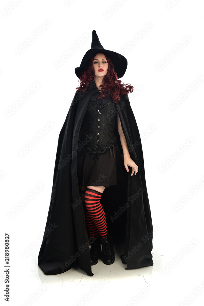 full length portrait of red haired girl wearing long black cloak, pointy hat and witch costume. standing pose, isolated on white studio background.