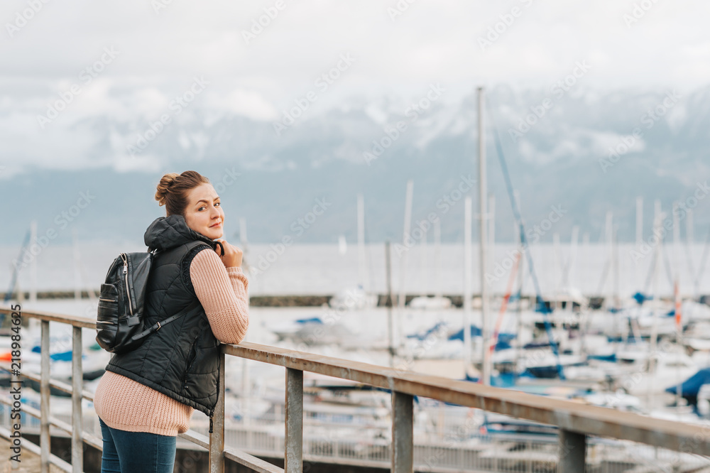 Outdoor portrait of young woman admiring winter lake, wearing warm pullover, black down vest and backpack. Image taken on Lake Geneva, Lausanne, Switzerland Outdoor portrait of young woman admiring winter lake, wearing warm pullover, black down vest and backpack. Image taken on Lake Geneva, Lausanne, Switzerland