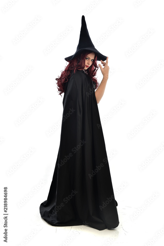 full length portrait of red haired girl wearing long black cloak, pointy hat and witch costume. standing pose, isolated on white studio background. full length portrait of red haired girl wearing long black cloak, pointy hat and witch costume. standing pose, isolated on white studio background.