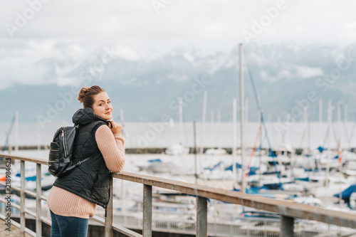 Outdoor portrait of young woman admiring winter lake, wearing warm pullover, black down vest and backpack. Image taken on Lake Geneva, Lausanne, Switzerland