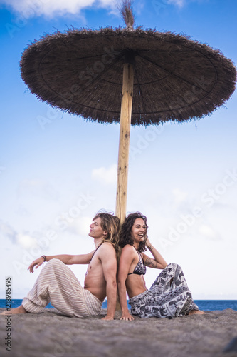 beautiful and cheerful caucasian young couple man woman sitting under an umbrella sun at the beach enjoying vacation and outdoor summer leisure activity. relax and love © simona