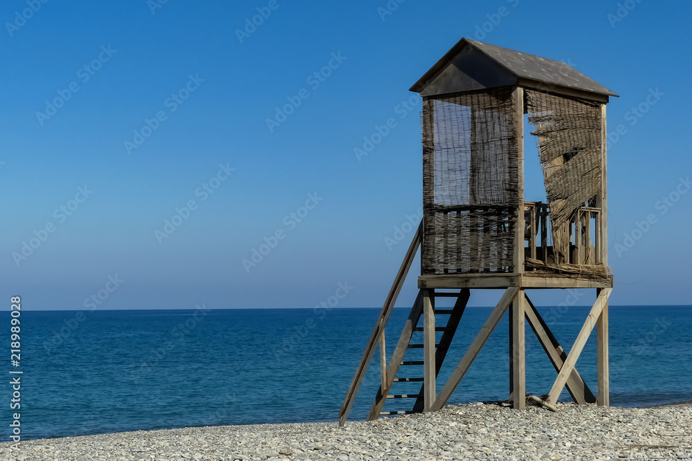 Abandoned rustic wooden lifeguard's tower on shingle beach, blue sky with sea horizon. Lightly rippling waves 