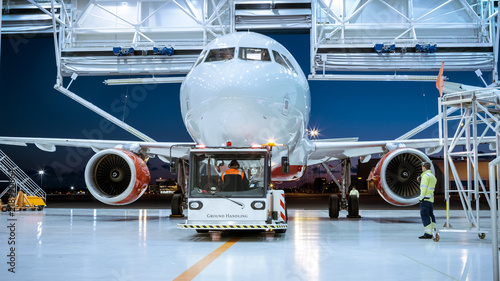 Aircraft Maintenance Hangar Where New Airplane is Toed by a Pushback Tractor/ Tug onto Landing Strip. Crew of Mechanics, Engineers and Drivers Works Busily.