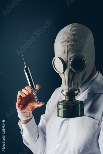 Creepy medical experiment concept, a scary doctor in gas mask wearing bloody gloves with a big stainless steel syringe on dark background