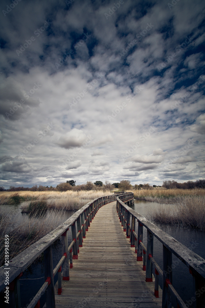 Obraz premium Puente de madera sobre laguna en un atardecer con nubes