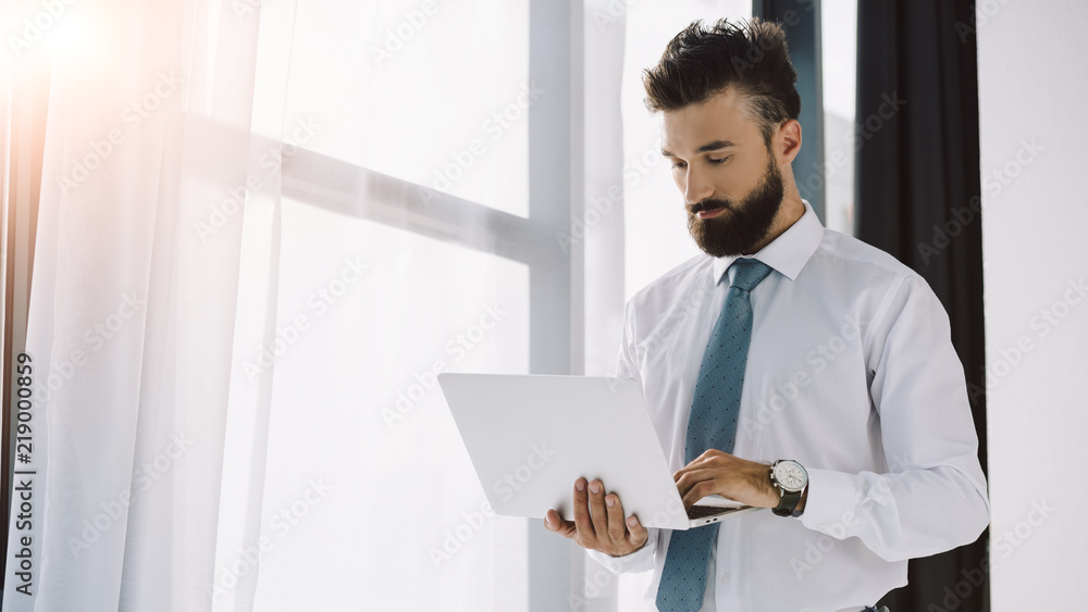 handsome bearded businessman using laptop near window in office handsome bearded businessman using laptop near window in office