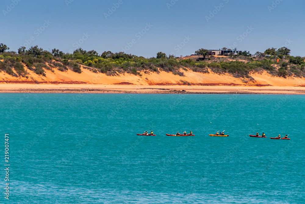 Broome, WA, Australia - November 29, 2009: Horizontal line made by red dunes with some green vegetation on top separates blue sky from azure water. Line of red canoes. Villa on top of one  dune.