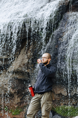Man drinks tea or coffee from a thermos, standing near a waterfall in the mountains