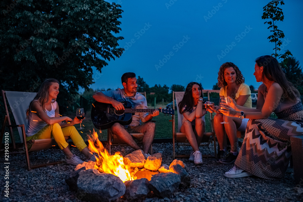 Enjoying the summer. Young man playing guitar for friends near bonfire at night. - obrazy, fototapety, plakaty 