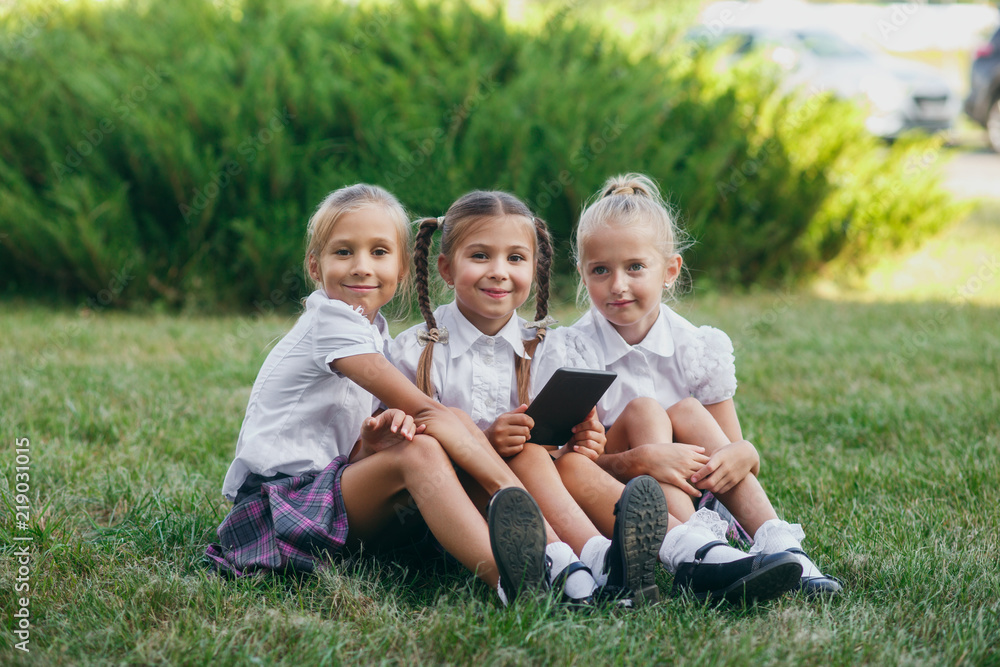Fototapeta premium three little girls are sitting on the grass and reading an e-book. Pupils of primary school. Beginning of lessons. First day of fall.